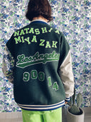 White woman with brown hair with her back to the camera wearing a color blocked letterman jacket with Custom chenille patches spelling Natashia Miyazaki Los Angeles 90014, the back is forest green, the right sleeve is a white lambskin leather with patches and the left sleeve is a custom printed velvet fabric. Royal blue & white stripe rib trim standing against a wallpaper that matches the print on the left sleeve. she also is wearing a neon green tracksuit under the jacket
